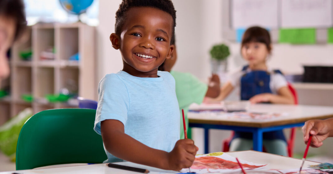 Young boy smiling while painting at a table in a classroom, with other children working in the background.