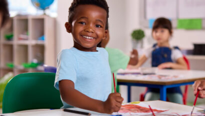 Young boy smiling while painting at a table in a classroom, with other children working in the background.