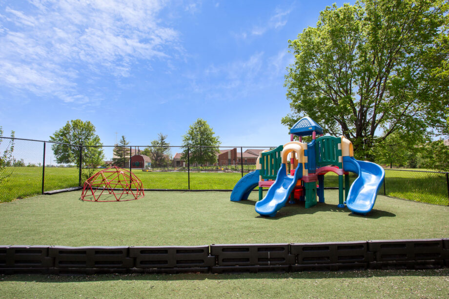A playground with slides, a climbing dome, and artificial turf is enclosed by a black fence with trees and buildings in the background under a blue sky.