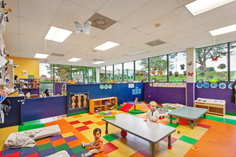 Daycare room with colorful flooring, low tables, mats, and young children playing; art and decorations on blue walls and large windows showing trees outside.