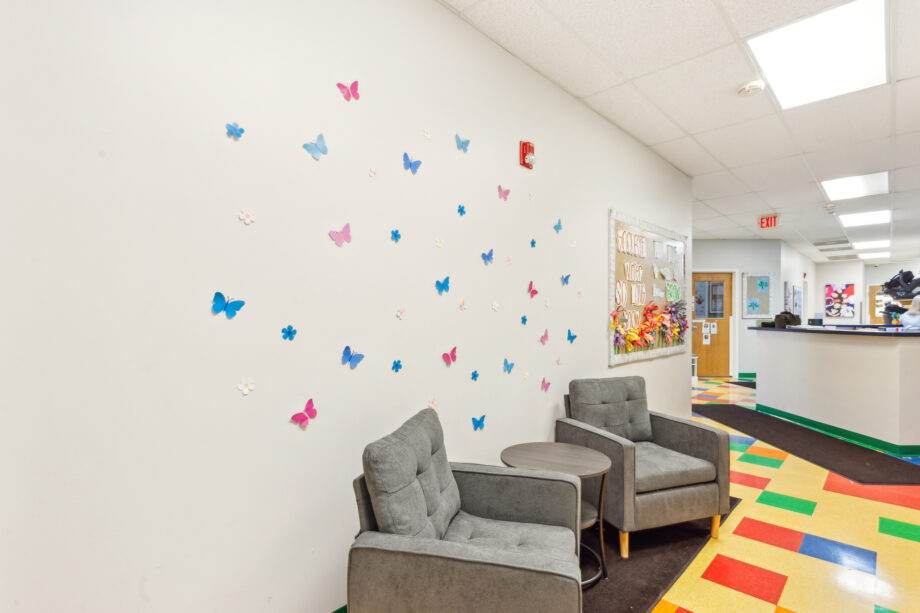 Two gray armchairs and a small table sit against a white wall decorated with colorful butterfly decals in a bright hallway with a multicolored tile floor.