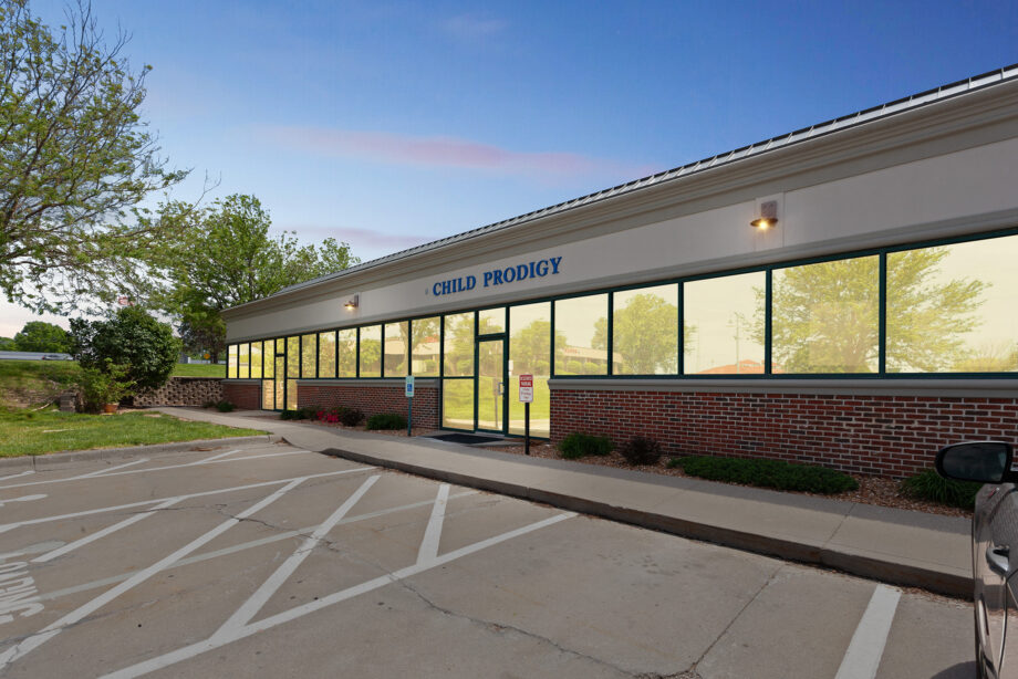Single-story building with "Child Prodigy" sign above glass entrance, surrounded by trees, with a parking lot featuring accessible parking spaces in front.