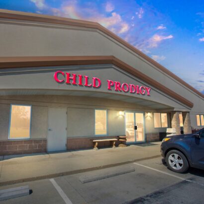 A building with a sign reading "Child Prodigy" above the entrance, illuminated at dusk, with a parked car in front.