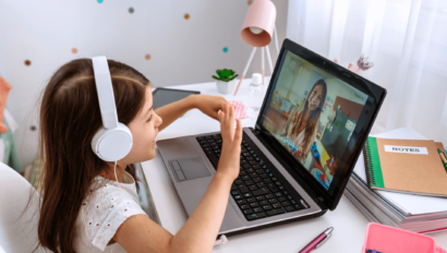 Young girl wearing headphones sits at a desk, waving at a woman on a laptop screen during a video call. Notebooks, a lamp, and other study materials are on the desk.