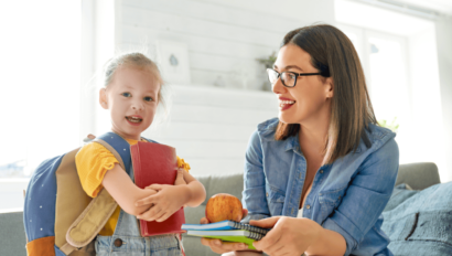 A young girl with a backpack holds books while an adult woman hands her more books and an apple, both smiling in a bright room.