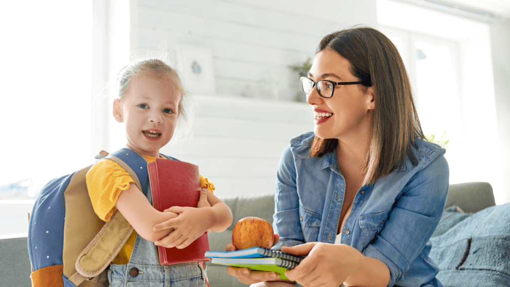 A young girl with a backpack holds books while an adult woman hands her more books and an apple, both smiling in a bright room.