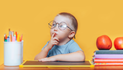 A young child wearing glasses sits at a desk with a finger on their lips, surrounded by books, apples, and a cup of pencils, against a plain yellow background.