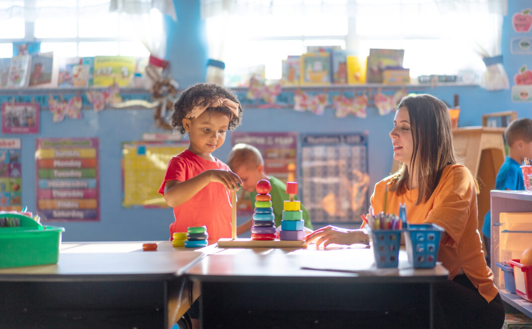 A young child stacks colorful rings while a teacher observes and writes at a desk in a brightly lit classroom.