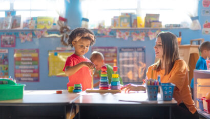 A young child stacks colorful rings while a teacher observes and writes at a desk in a brightly lit classroom.