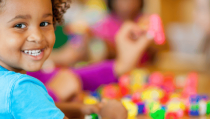 A young child smiles at the camera while playing with colorful building blocks at a table. Other children are blurred in the background.