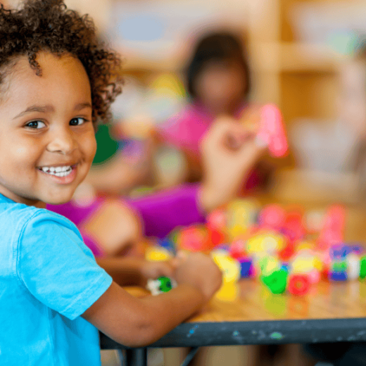 A young child smiles at the camera while playing with colorful building blocks at a table. Other children are blurred in the background.