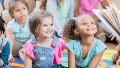 Two young girls sitting on a colorful mat, smiling and looking toward an adult reading a book, with other children seated in the background.
