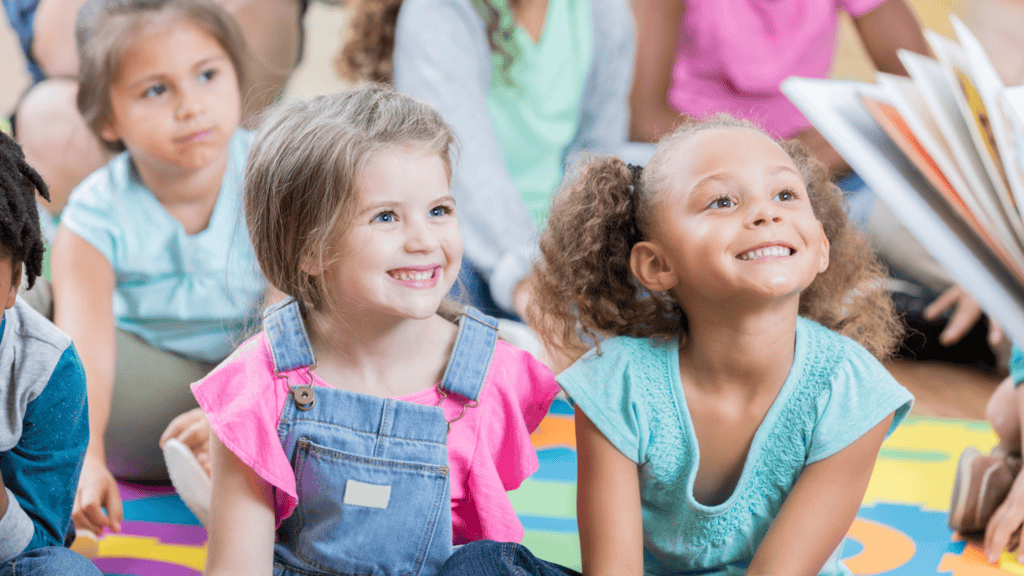 Two young girls sitting on a colorful mat, smiling and looking toward an adult reading a book, with other children seated in the background.