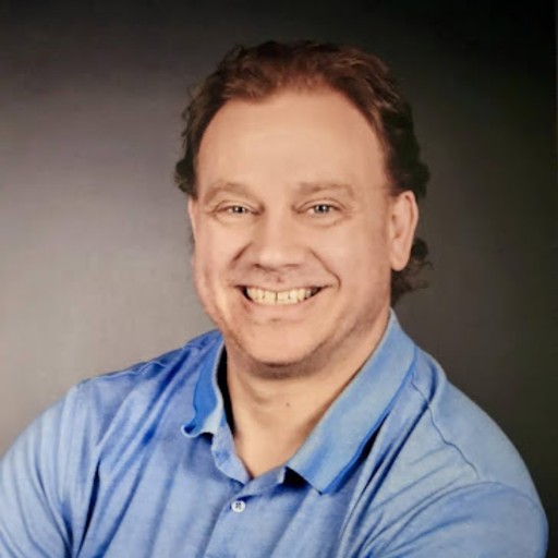 A man with light skin and short brown hair smiles at the camera while wearing a light blue collared shirt, posed against a dark background.