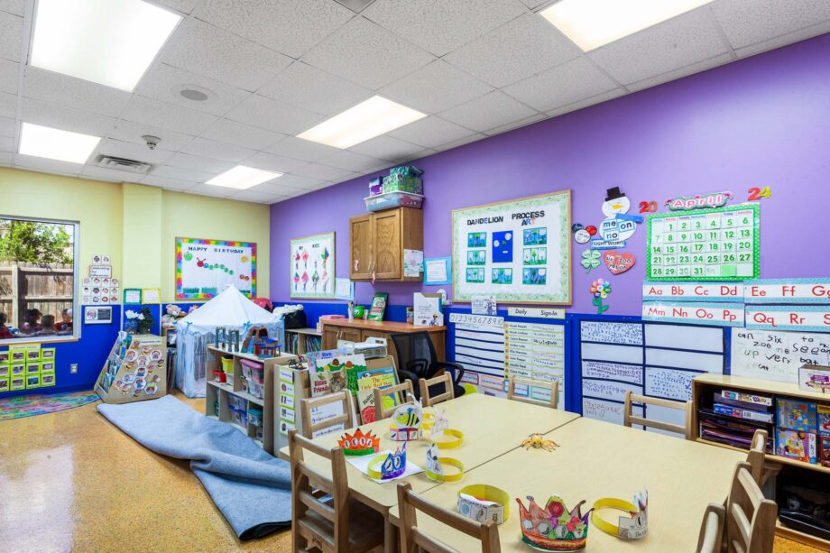 A brightly lit preschool classroom with colorful decorations, child-sized tables and chairs, educational posters, and various learning materials organized on shelves.