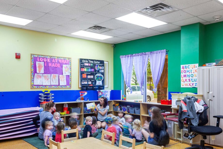 A group of young children sit on the floor in a classroom while two adults read to them; colorful posters and toys are visible around the room.
