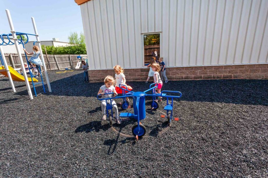 Children play on a blue merry-go-round and climbing equipment on a playground with a black rubber surface next to a white building.