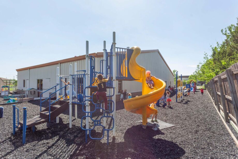 Children play on a playground with climbing structures and a yellow spiral slide on a sunny day, with a building and trees in the background.