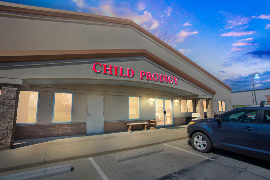 A building with a sign reading "CHILD PRODIGY" above the entrance, with a car parked in front and the sky at dusk in the background.