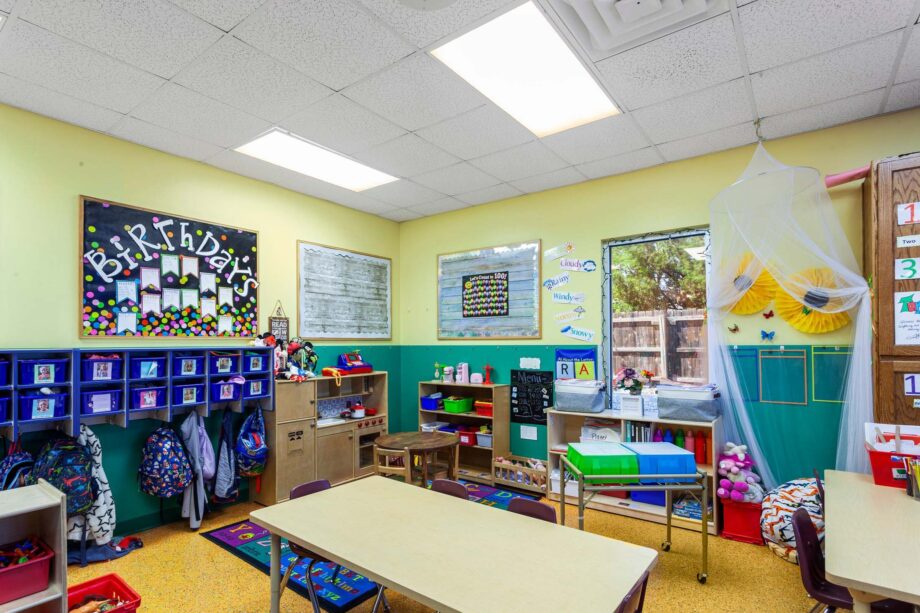A brightly lit classroom with small tables, chairs, cubbies with backpacks, colorful wall decorations, and a "Birthdays" bulletin board.